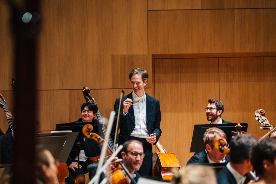 ein lachender Dirigent im schwarzen Frack dirigiert ein Symphonieorchester